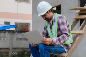 A construction project manager is sitting on partially constructed steps using his tablet to check the potential tax implications of purchasing new equipment